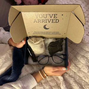 Open cardboard box with 'You've Arrived' text, containing baby items on a textured surface.